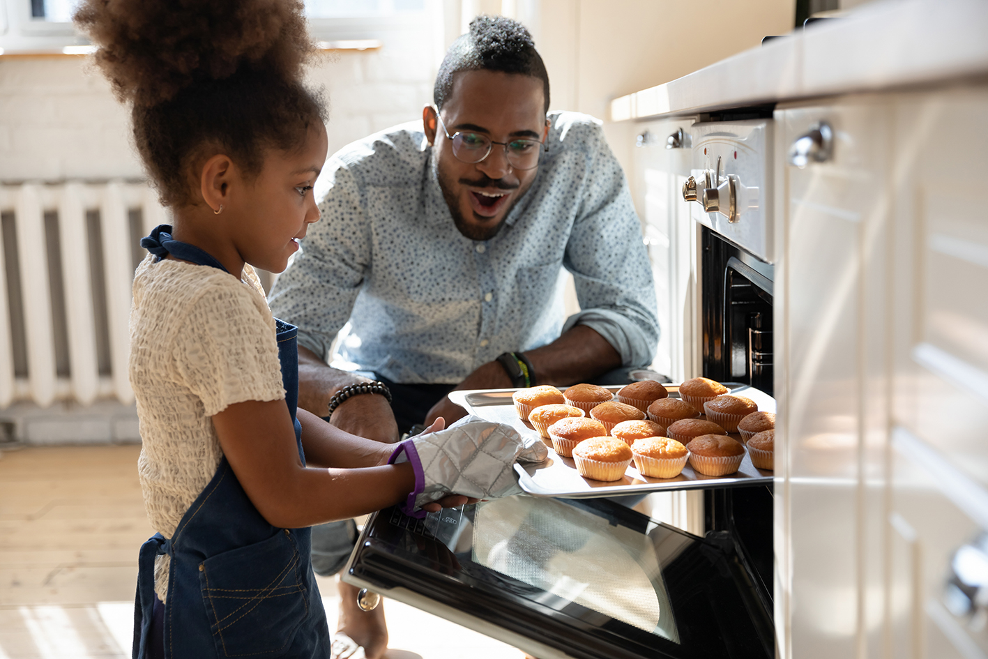 child takes buns out of oven with parent crouched down next to them with a smile on their face